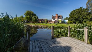 A wooden dock leading to a calm water body with a grassy area and a large building in the background. - The Moat House (Stafford)