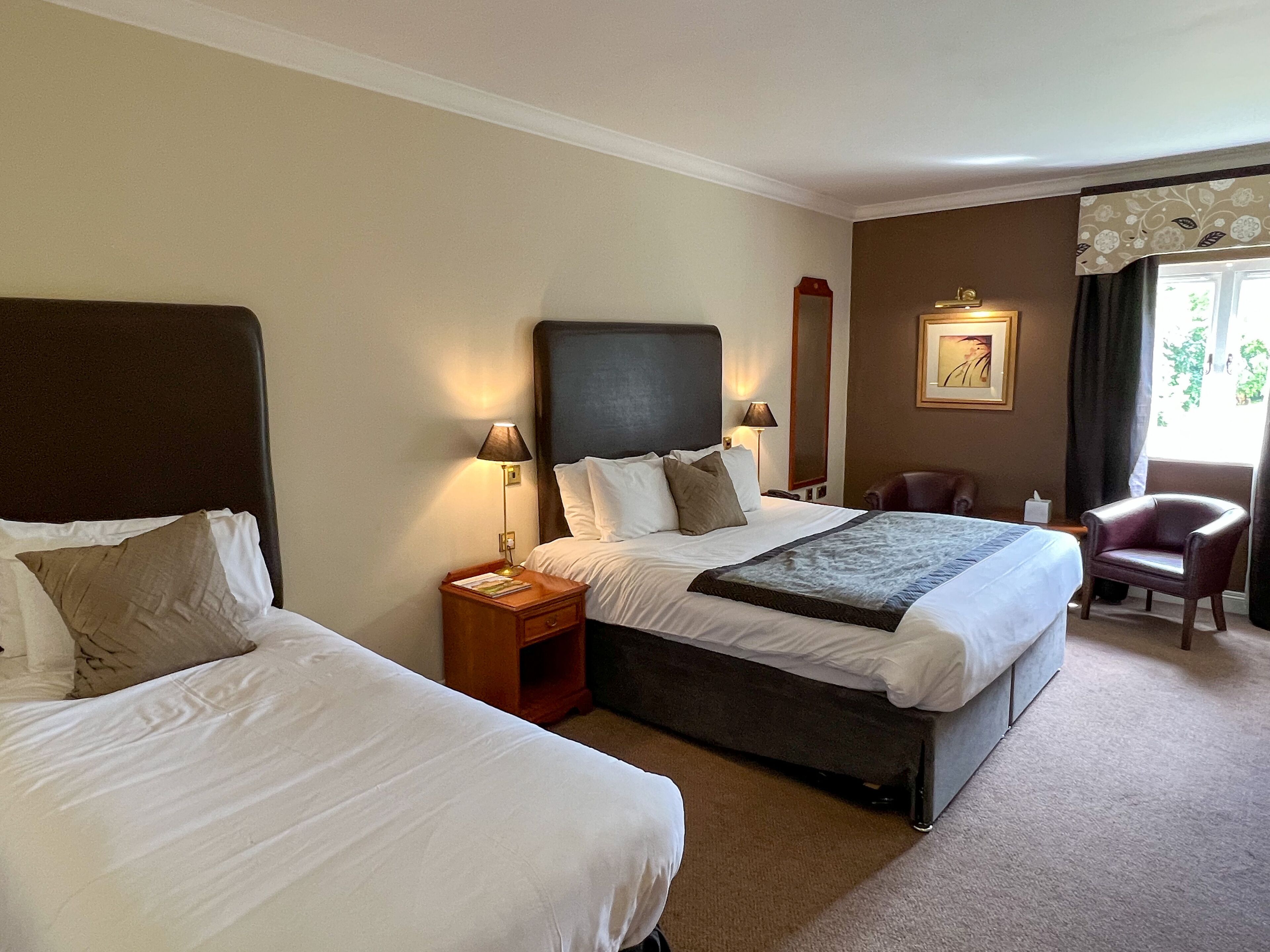 A hotel room with two beds, a nightstand with a lamp, a chair, a mirror, and a window with curtains.