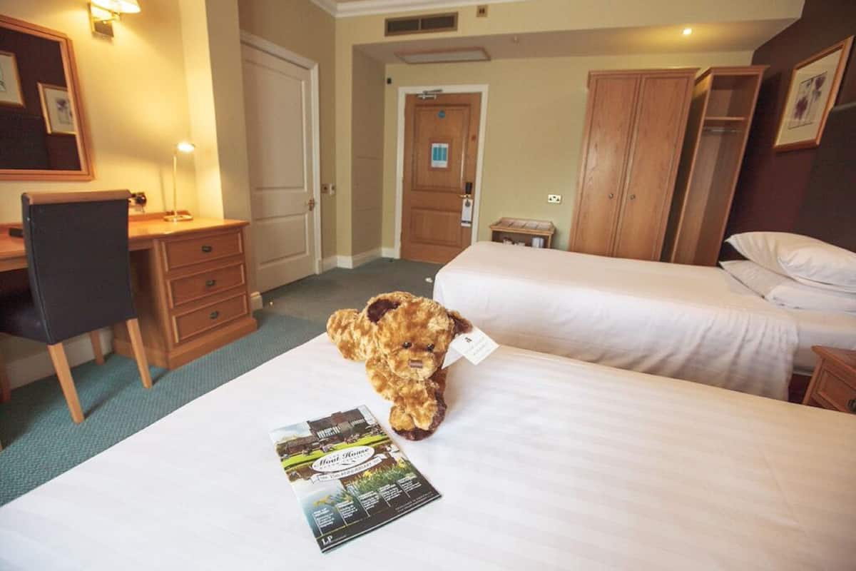 A hotel room with two beds, a desk, a chair, a teddy bear, and a brochure on the bed.