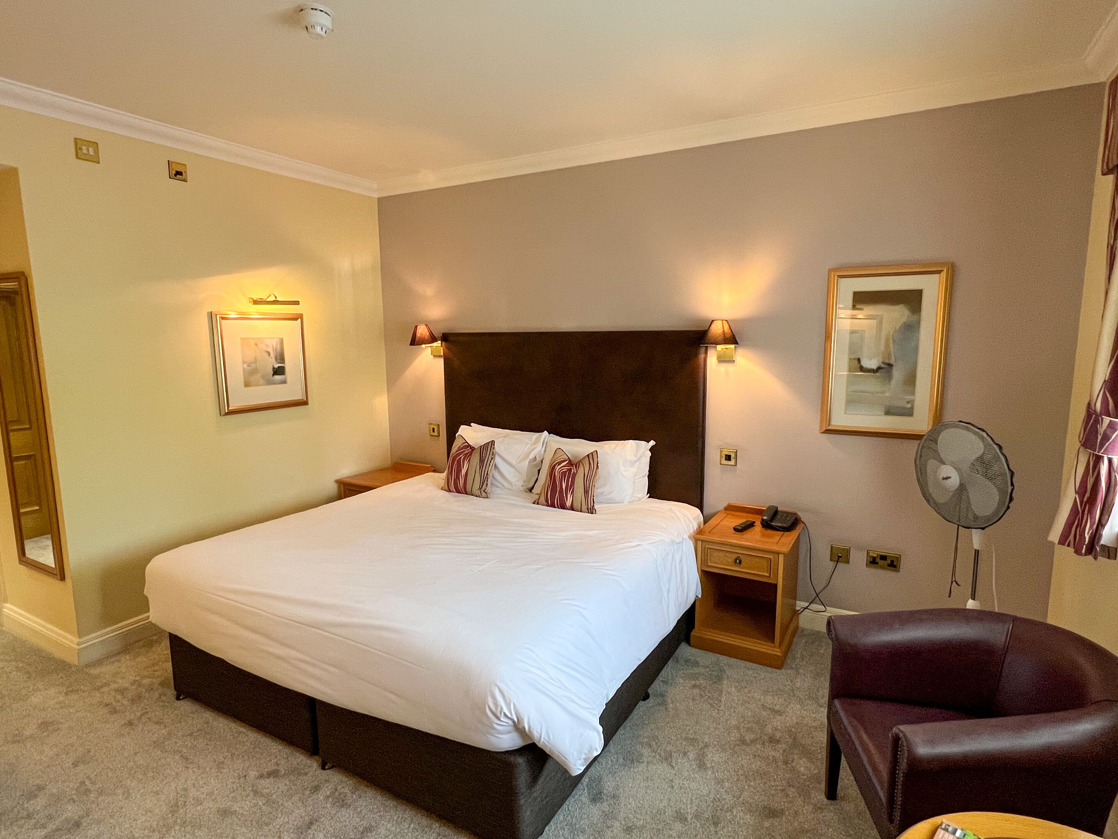 A hotel room with a large bed, bedside lamps, a desk with a telephone, a chair, and a standing fan.
