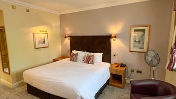 A hotel room with a large bed, bedside lamps, a desk with a telephone, a chair, and a standing fan.