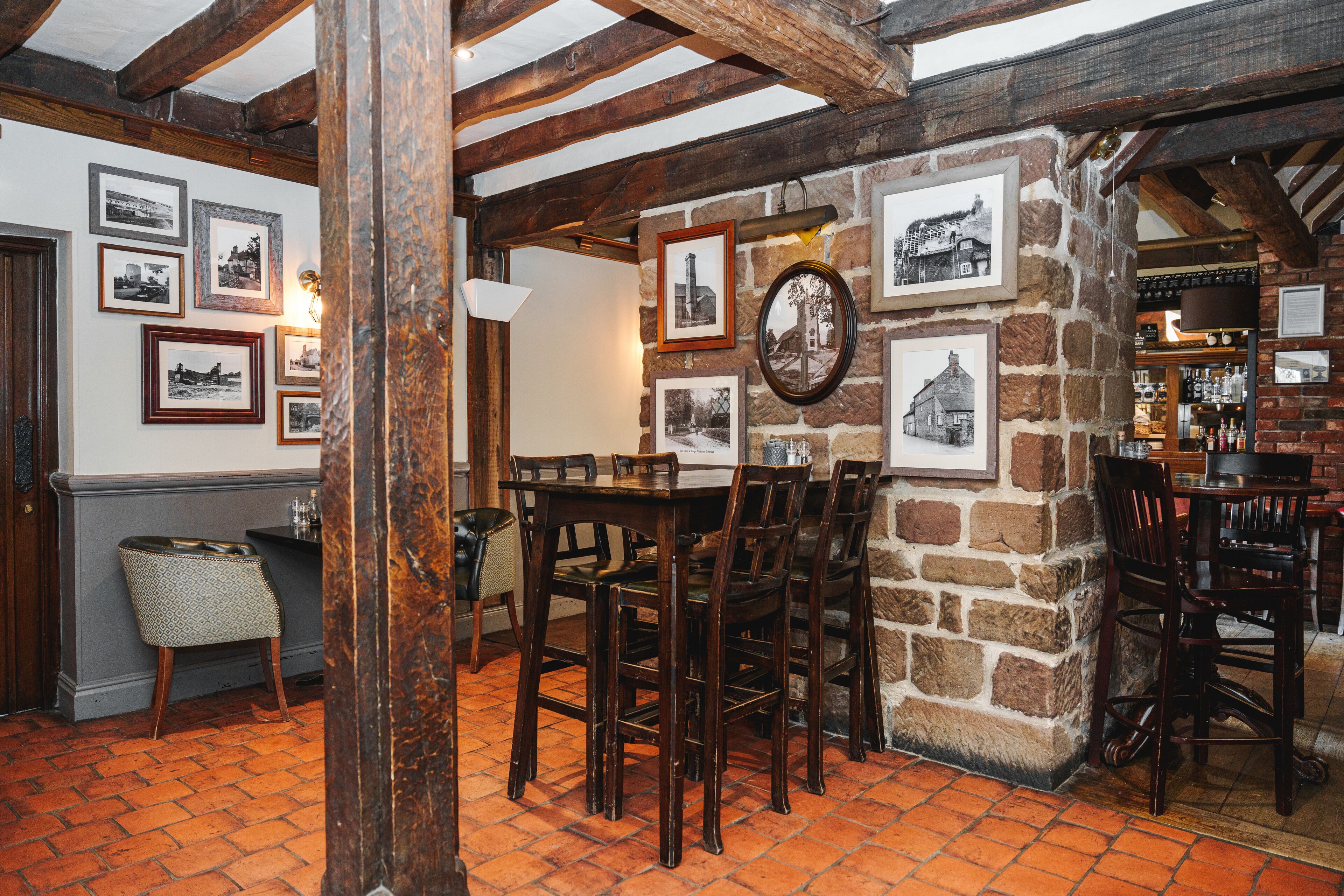 A rustic interior with a stone wall, wooden beams, and a brick floor. There are framed pictures on the wall and a bar area with bottles.