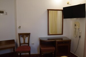 A room with a wooden chair, a desk with a mirror, a television mounted on the wall, and a telephone.