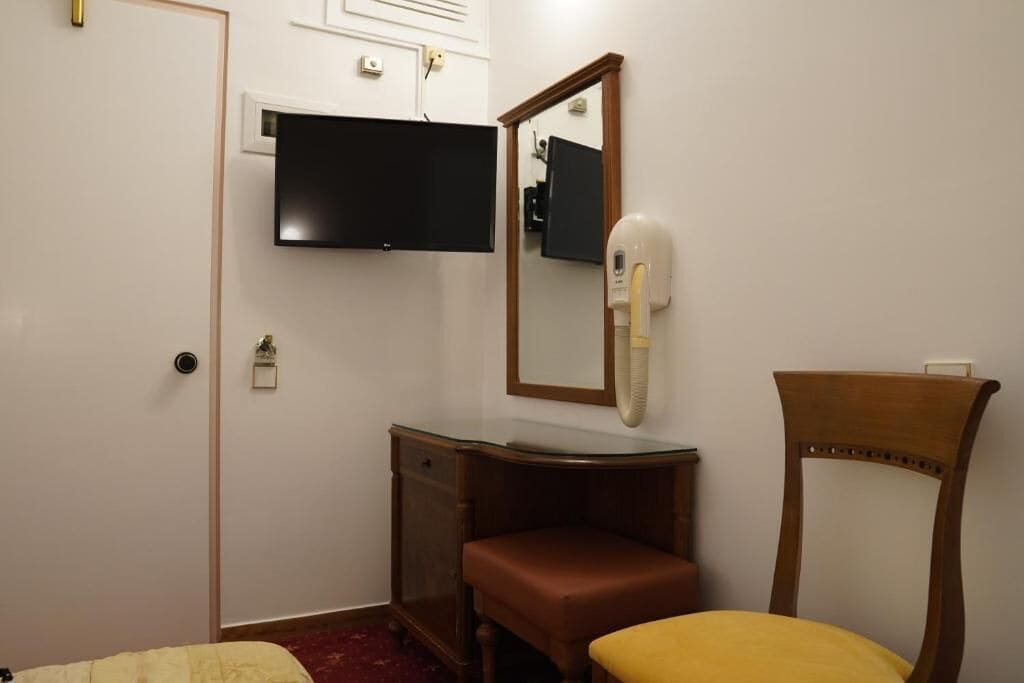 Ein Zimmer mit einem Bett, einem Holzstuhl, einem Schminktisch mit Spiegel und einem an der Wand befestigten Fernseher.