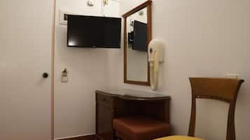 Ein Zimmer mit einem Bett, einem Holzstuhl, einem Schminktisch mit Spiegel und einem an der Wand befestigten Fernseher.