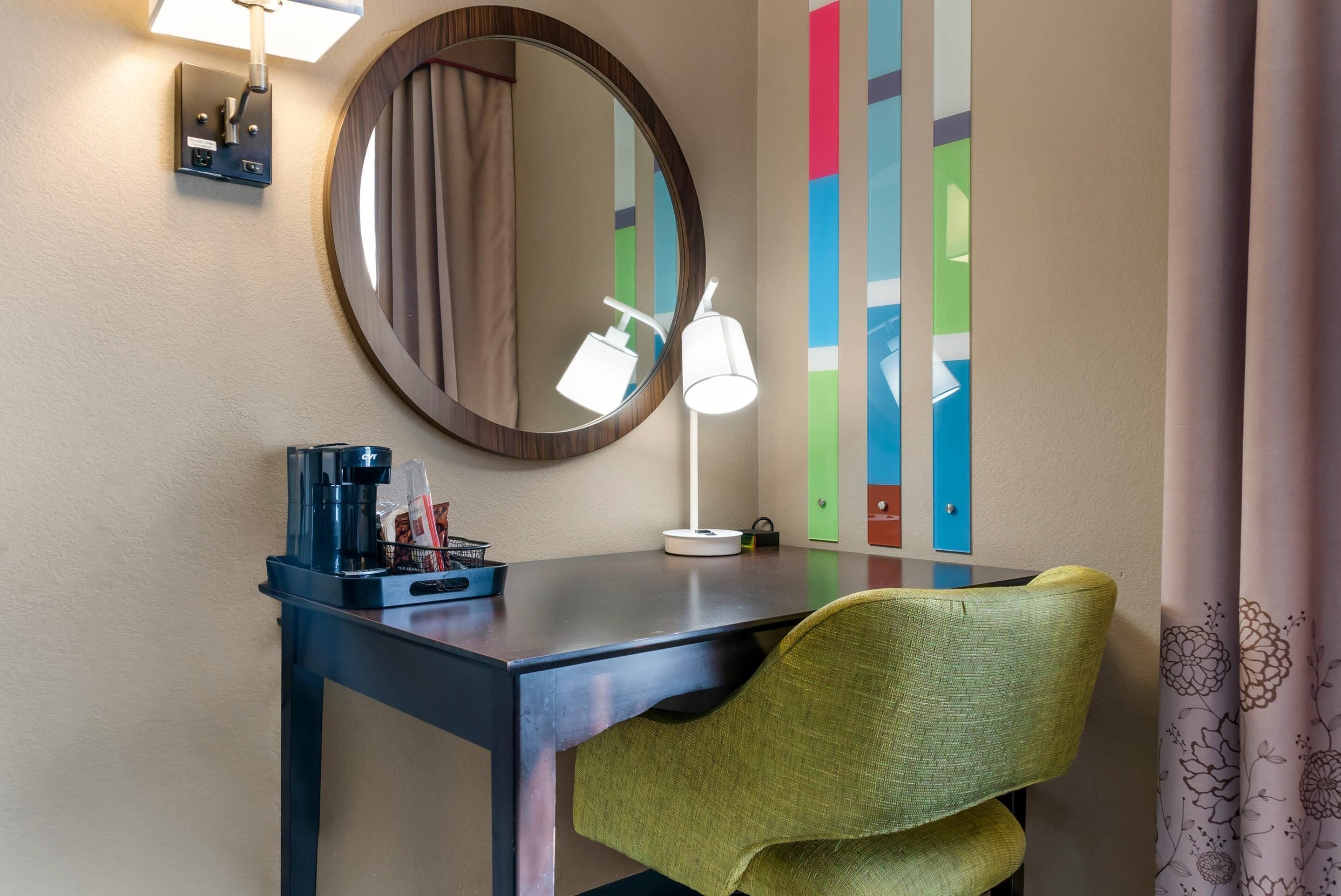 Una habitación de hotel con escritorio, una silla, cafetera, lámpara, espejo y un colorido adorno de pared.