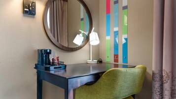 Una habitación de hotel con escritorio, una silla, cafetera, lámpara, espejo y un colorido adorno de pared.