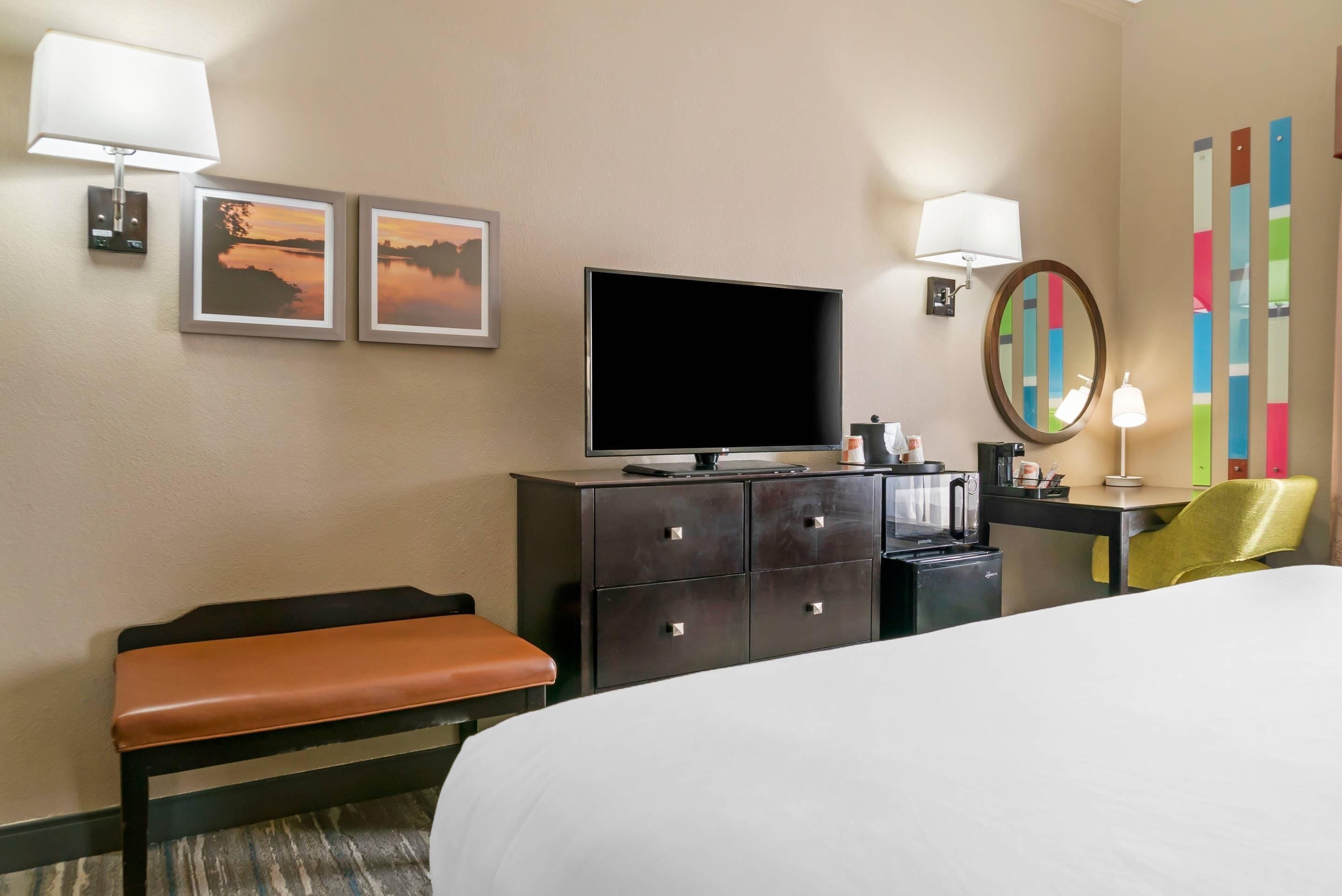 A hotel room with a bed, a flat-screen TV, a dresser, a bench, a desk with a lamp, a mirror, and two framed pictures on the wall.