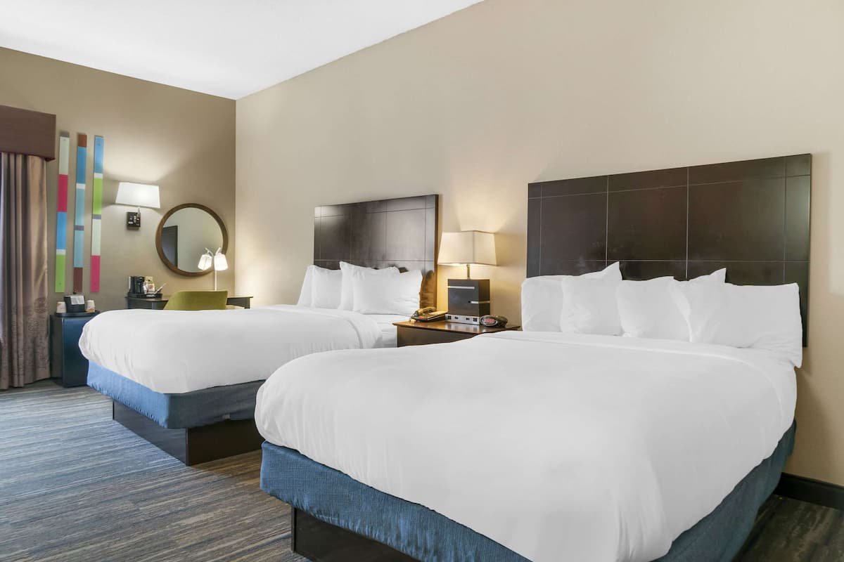 Habitación de hotel con dos camas, un espejo grande, una mesita de noche con lámpara y una pared con un diseño abstracto y colorido.