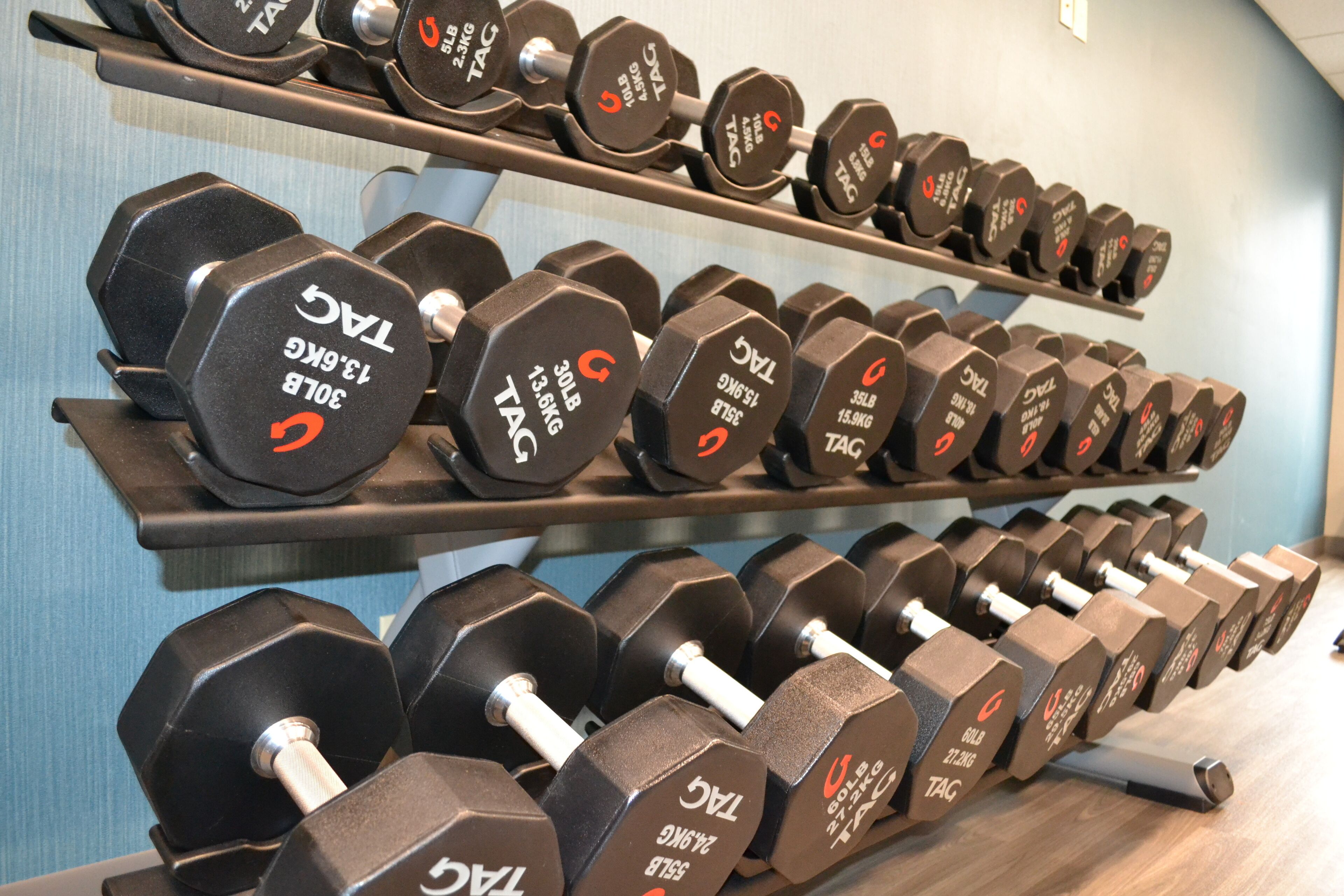 A rack of dumbbells with weight labels, arranged on shelves in a gym setting.