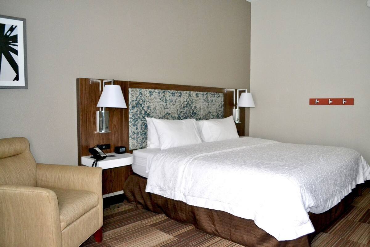 A hotel room with a bed, bedside lamps, a nightstand, a chair, and a wall clock.