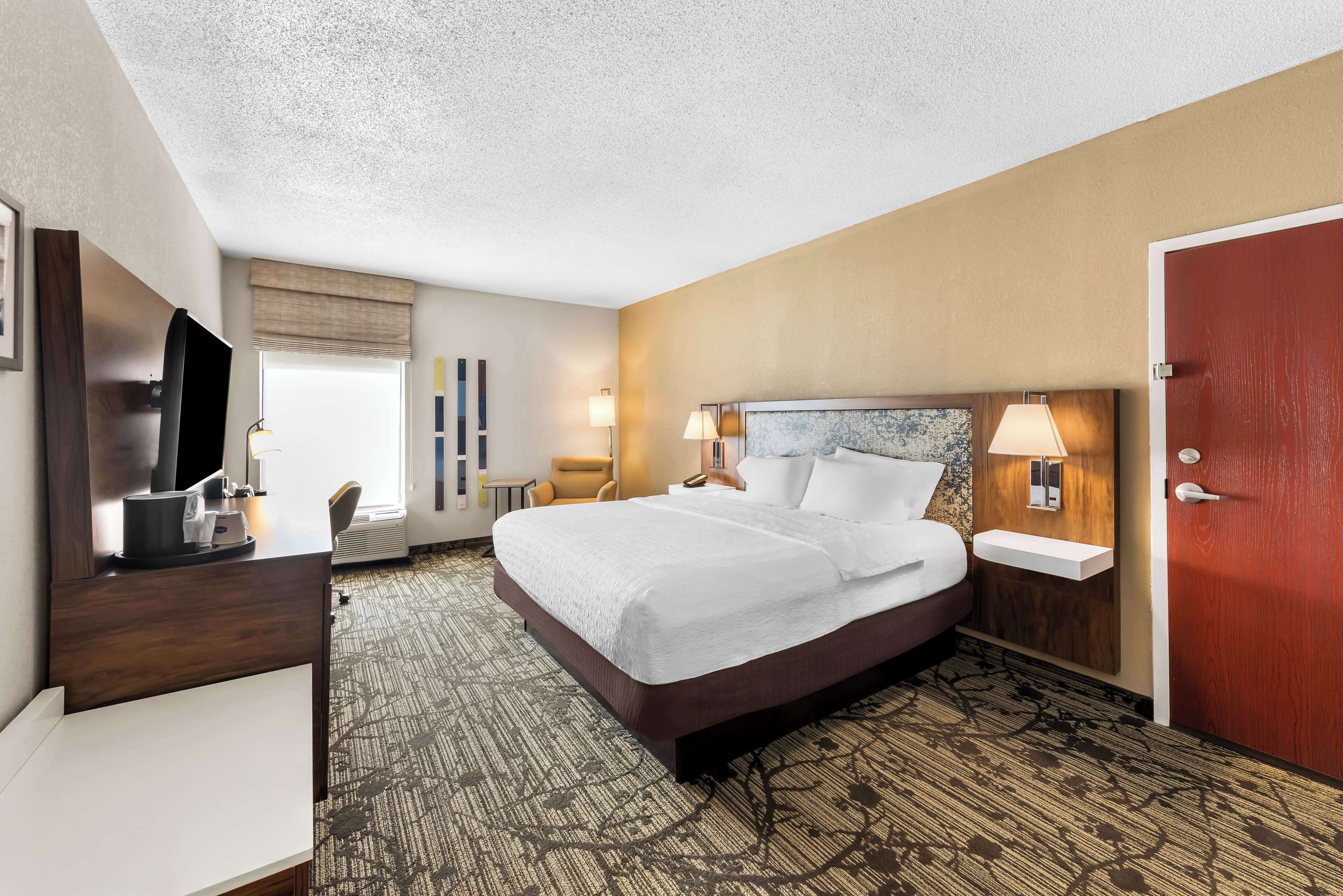 A hotel room with a large bed, a desk with a flat-screen TV, a chair, and a red door.