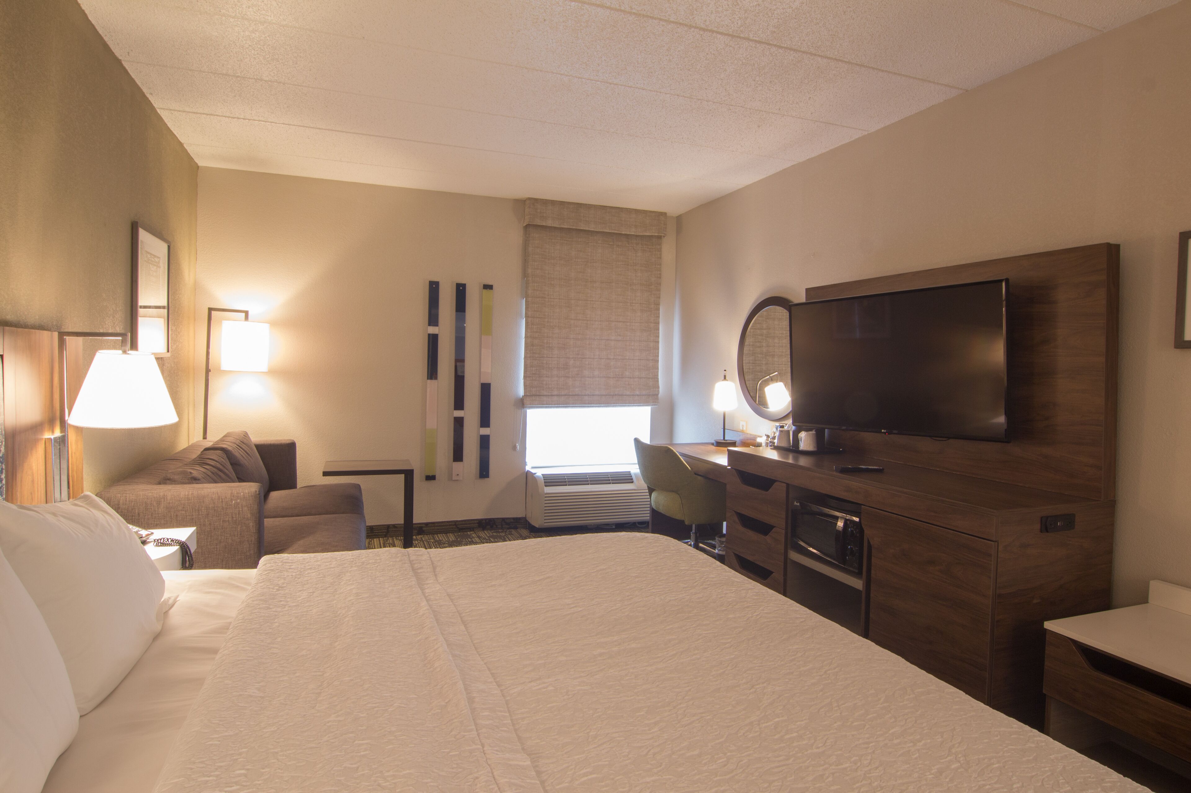 A hotel room with a large bed, a desk with a flat-screen TV, a chair, a desk lamp, a mirror, and a window with curtains.