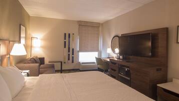 A hotel room with a large bed, a desk with a flat-screen TV, a chair, a desk lamp, a mirror, and a window with curtains.