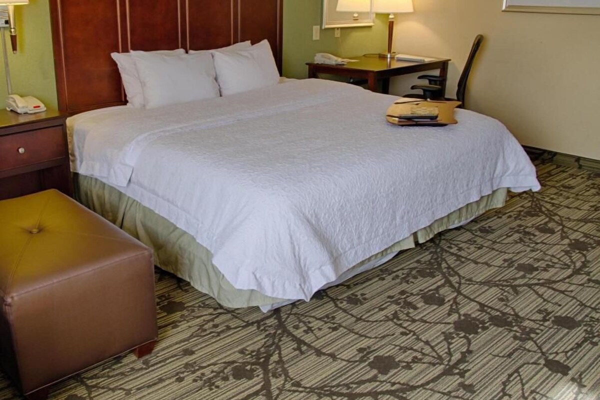 A hotel room with a large bed, a nightstand with a phone, a desk with a lamp, and a chair.