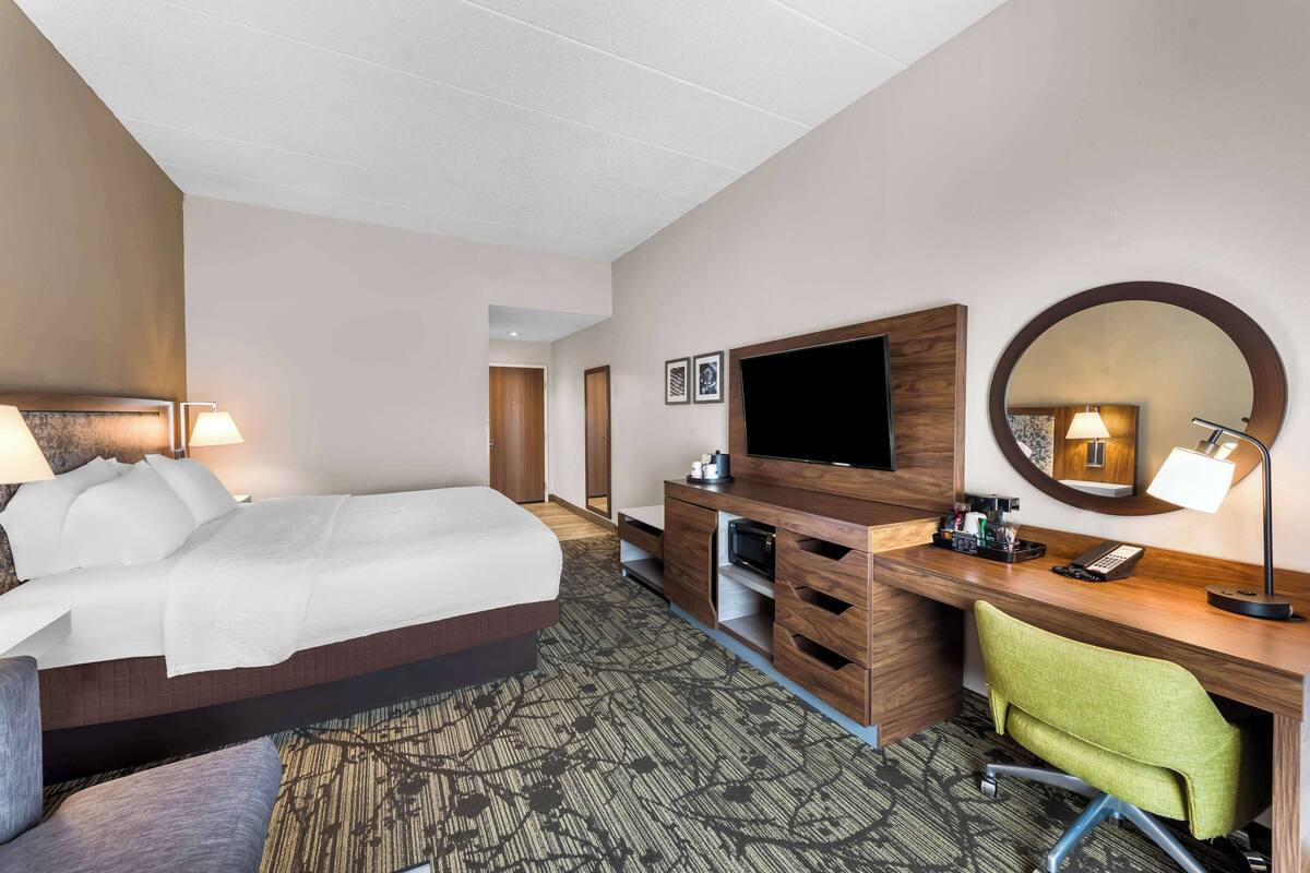 A hotel room with a bed, a desk with a computer, a TV, a mirror, and a chair.
