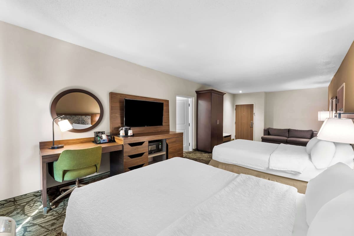 A hotel room with two beds, a desk with a TV, a green chair, a microwave, and a wardrobe.