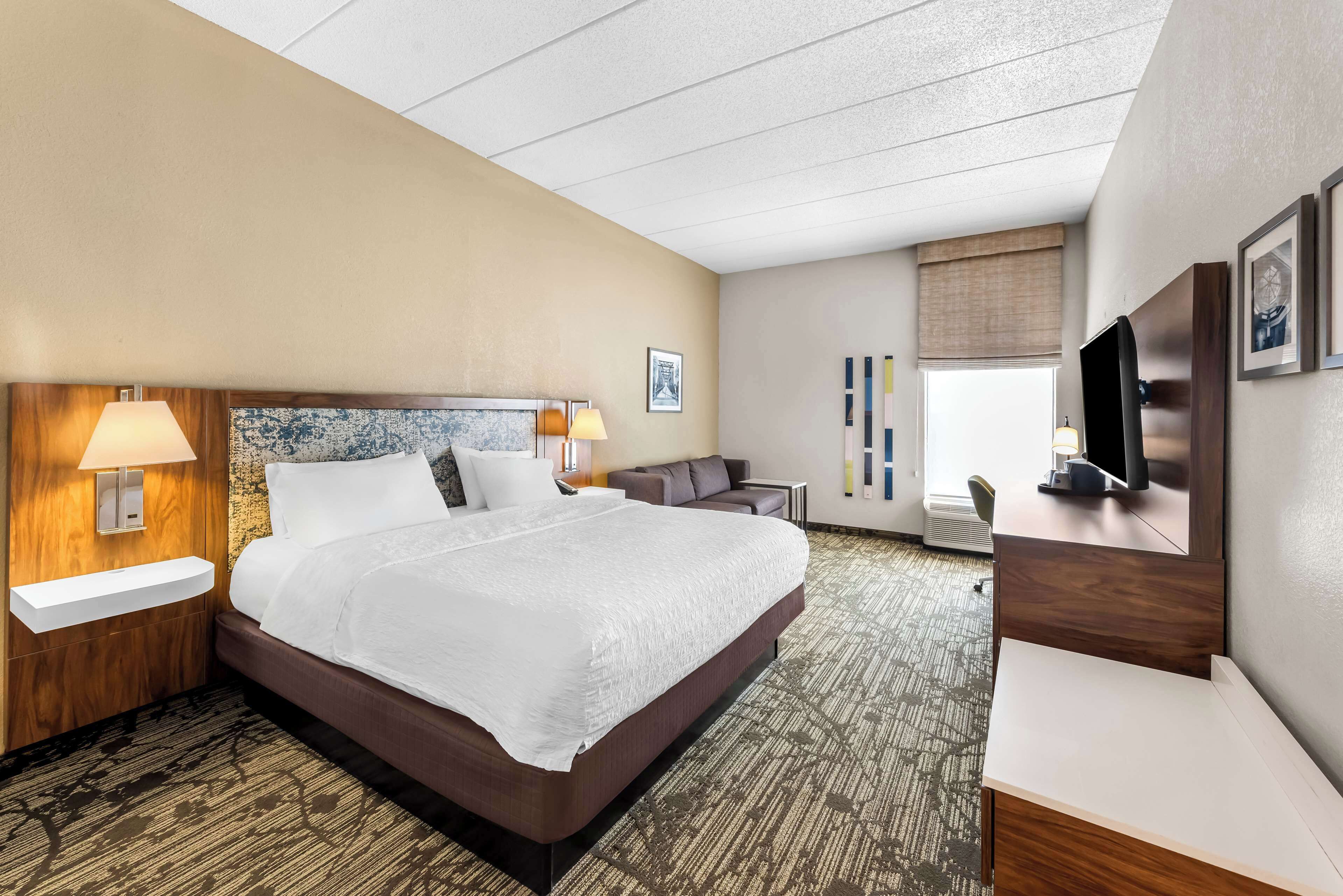 Ein Hotelzimmer mit einem großen Bett, Nachttischlampen, einem Schreibtisch mit Flachbildfernseher, einem Sofa und einem Fenster mit Jalousien.
