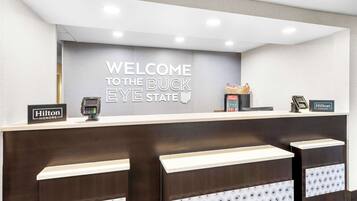 Una recepción de hotel con un letrero que dice "Welcome to the Buck Eye State", un cartel de Hilton Honors, un quiosco de autoservicio y una caja registradora.