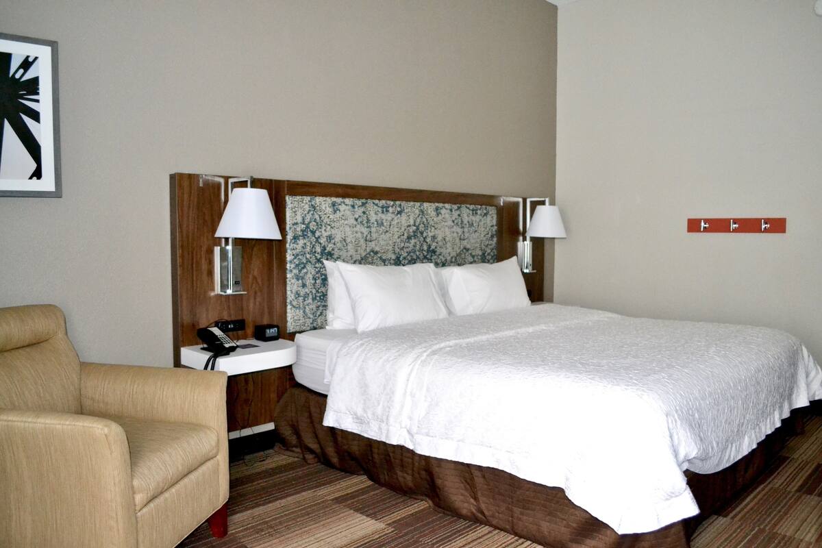 A hotel room with a large bed, two bedside lamps, a chair, a bedside table with a phone, and a wall-mounted control panel.