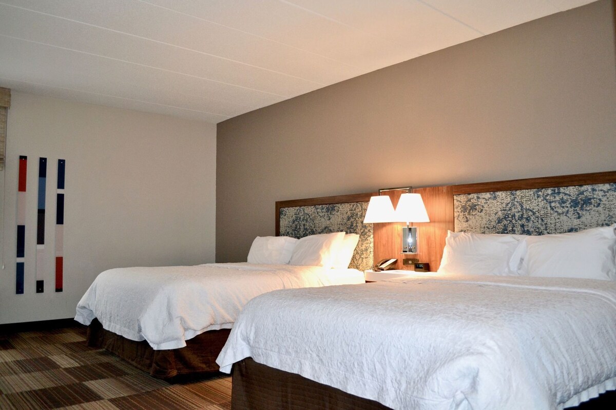 A hotel room with two beds, a wooden headboard, a bedside table with a lamp, and a wall with vertical stripes.