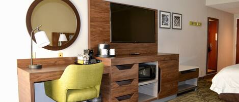 A hotel room with a wooden desk, a green chair, a flat-screen TV, a mirror, and a bed.