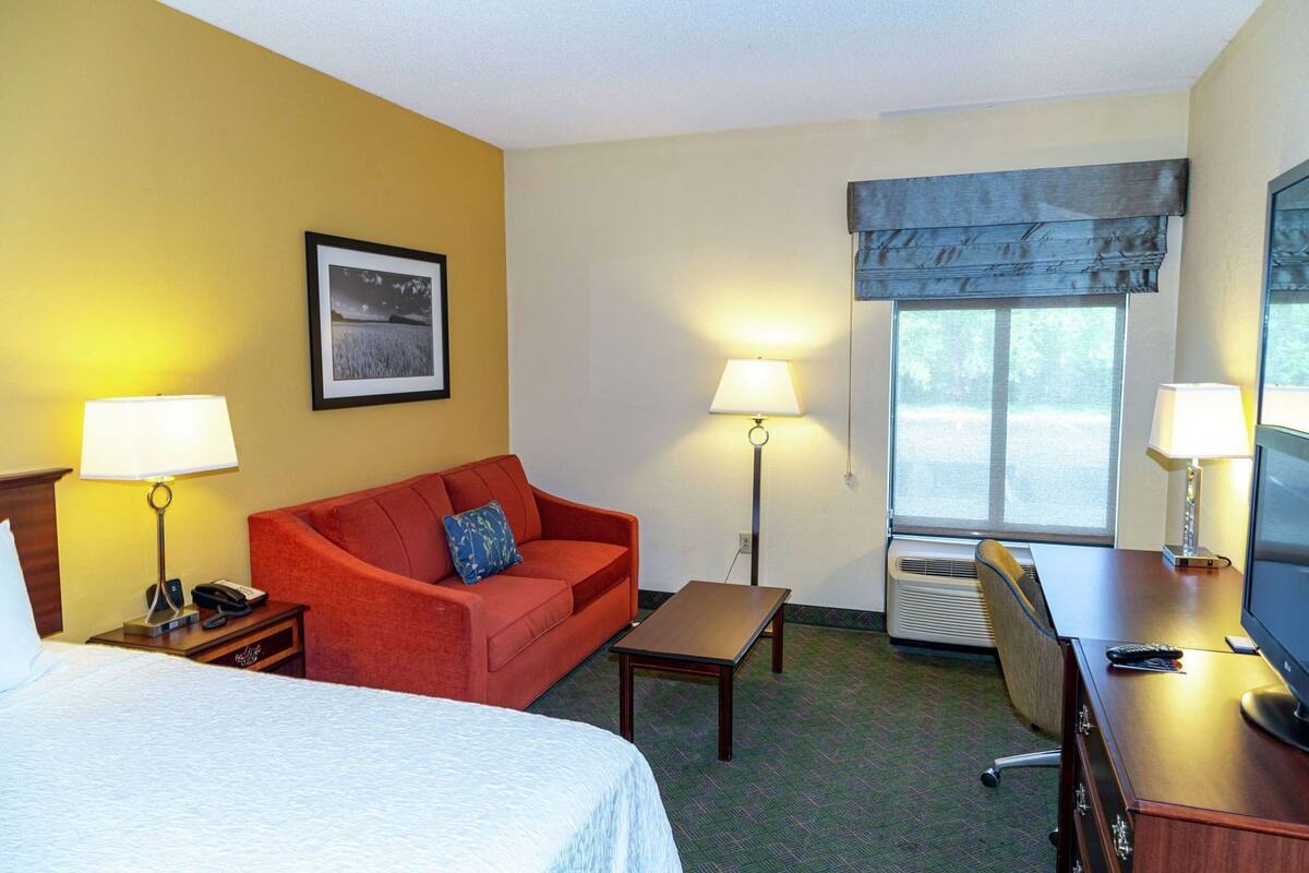 A hotel room with a bed, a red sofa, a small table, a desk with a chair, a TV, and a window with a view.