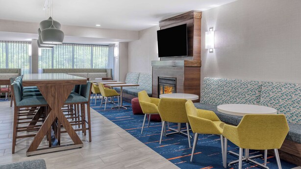 A modern hotel lobby with a wooden bar, yellow chairs, a fireplace, and a large TV on the wall.
