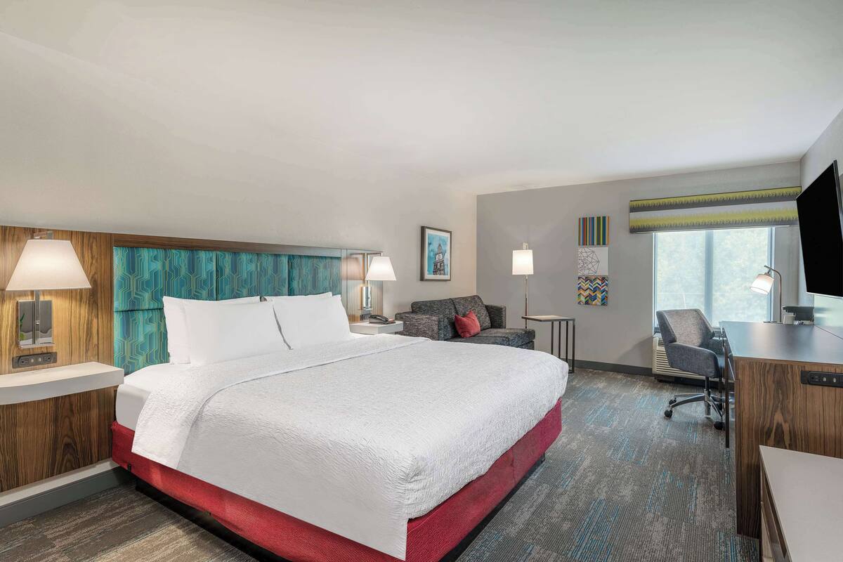 A hotel room with a large bed, a desk, a chair, a TV, and a window.