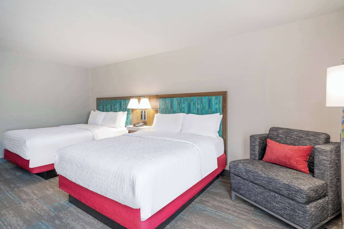 A hotel room with two single beds, a grey armchair, and a nightstand with a lamp.