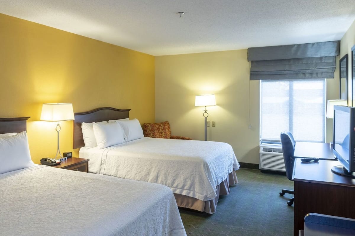 A hotel room with two beds, bedside lamps, a desk with a computer, and a window with a view.