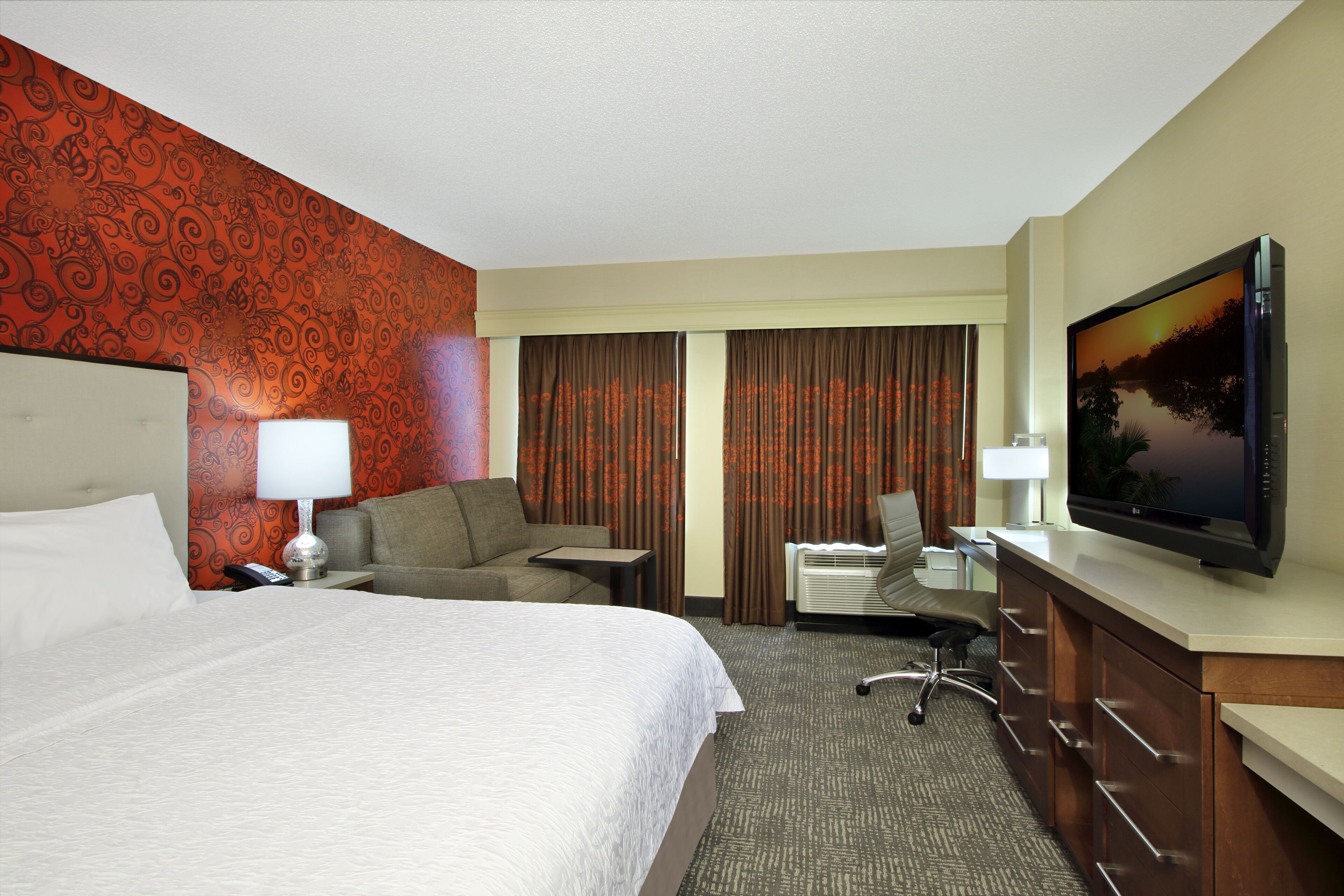 Une chambre d’hôtel dotée d’un grand lit, d’un bureau avec un ordinateur, d’une télévision et d’un coin salon comprenant un canapé et des fauteuils.
