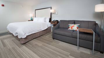 A hotel room with a bed, a gray sofa, a small table, and a nightstand with a lamp.
