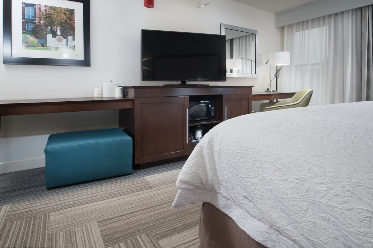Hotellihuone, jossa on sänky, televisio, työpöytä tuolilla, mikroaaltouuni ja seinällä kehystetty kuva.