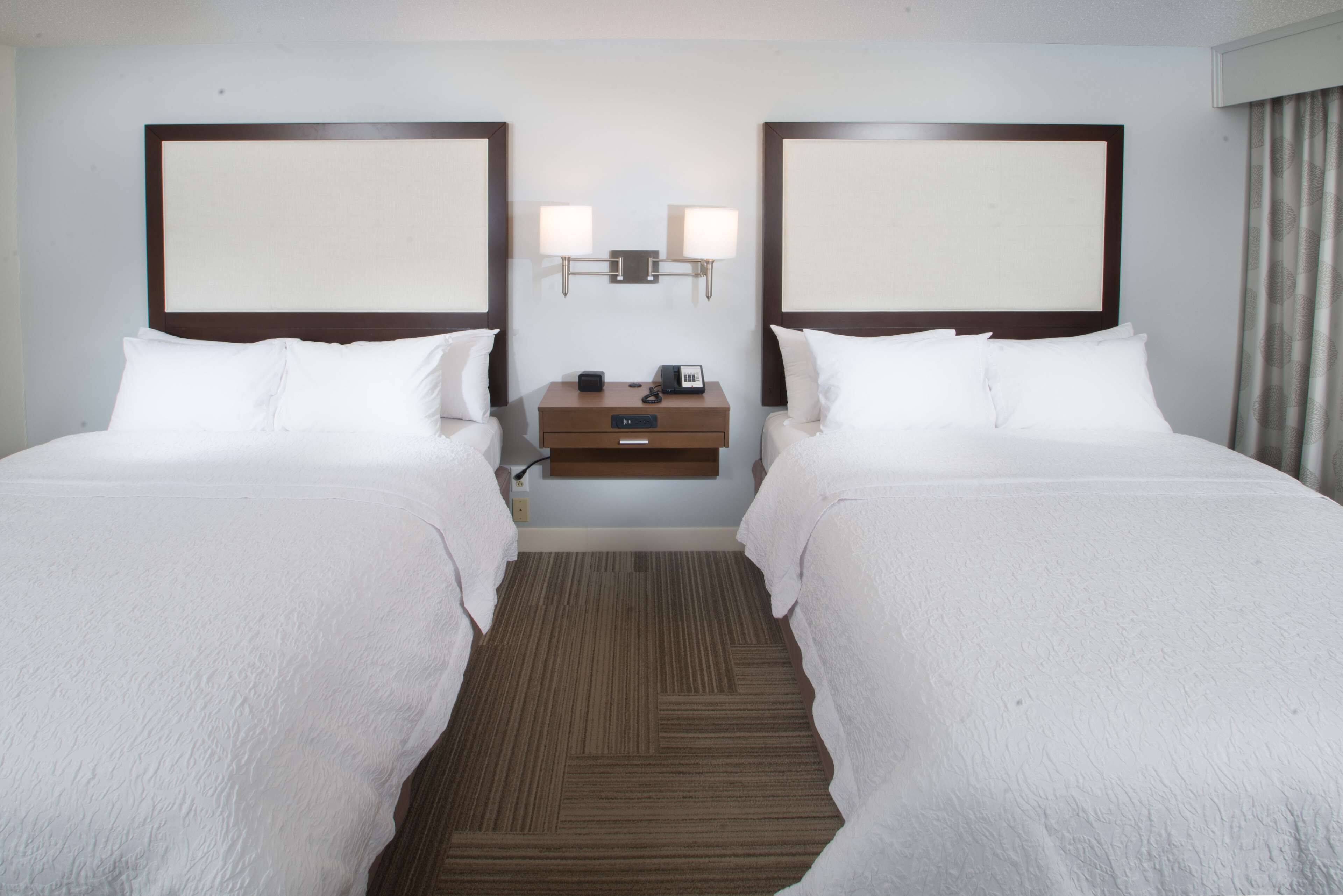 A hotel room with two beds, a nightstand, a lamp, and a wall-mounted light fixture.