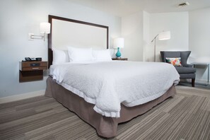 A hotel room with a large bed, a nightstand, a desk lamp, a chair, and a wall-mounted lamp.