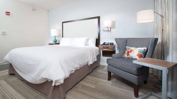 A hotel room with a large bed, a grey armchair, a small table, and a nightstand with a lamp.
