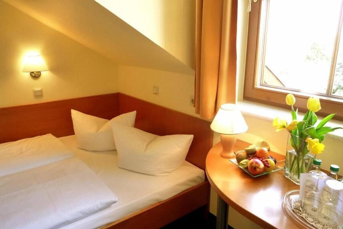 Une chambre d’hôtel avec un lit bien fait, une table de chevet avec une lampe, un vase rempli de fleurs et une assiette de fruits.