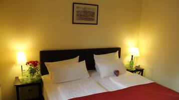 A neatly made bed with white linens and a red blanket, two bedside lamps, a small table with a vase of flowers, and a framed picture on the wall.