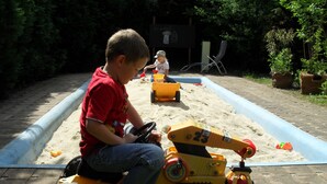 A child on a toy vehicle in a sandy play area with a blue water feature and a chair in the background. - Landhotel Dresden (Dresden)