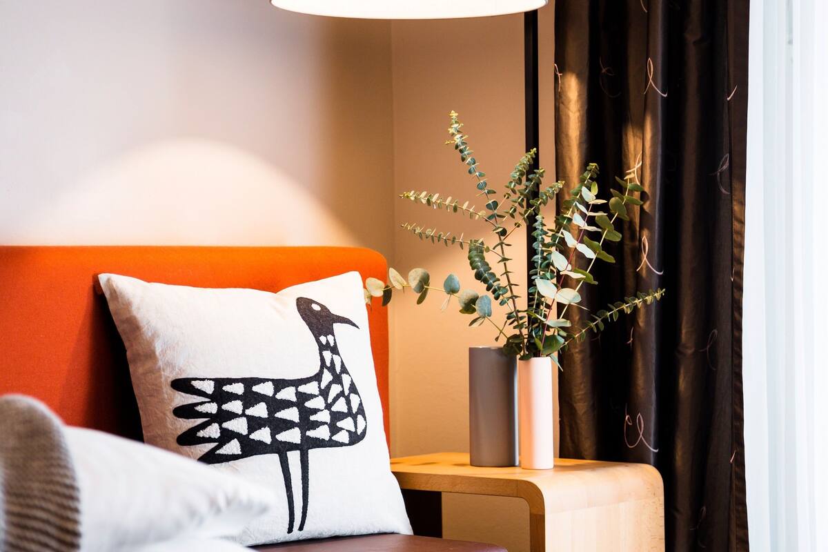 Una habitación de hotel con cama, mesita de noche, lámpara, planta en maceta y ventana con cortinas.