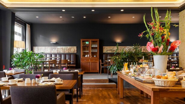 Un restaurant moderne avec des tables et des chaises en bois, un buffet proposant une variété de plats et un grand vase rempli de fleurs.