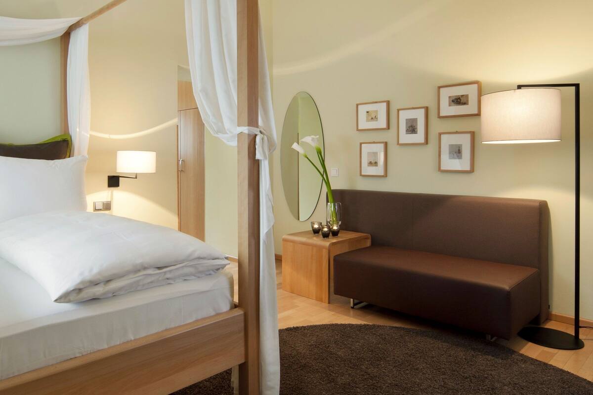 Habitación de hotel con cama con dosel, sofá, mesita de noche con lámpara y cuadros en la pared.
