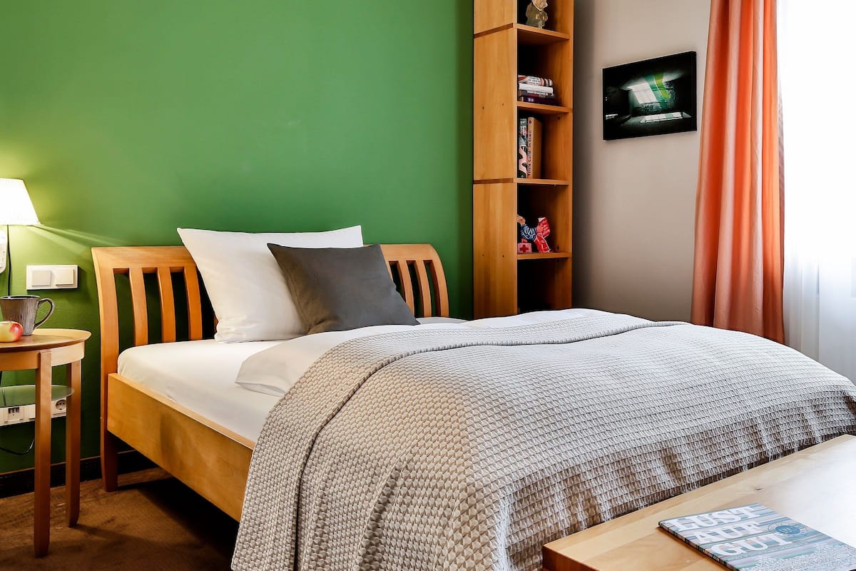 Una cama bien hecha con una colcha estampada, una mesita de noche con una lámpara, una pequeña mesa con un libro y una estantería con libros y un marco de fotos.