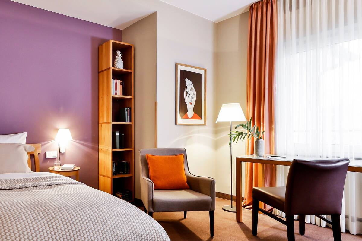 Una habitación de hotel con una cama, una estantería, una silla, un escritorio y una ventana con cortinas.