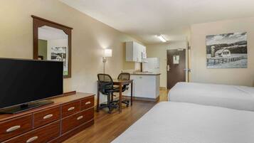 A hotel room with a TV, wooden dresser, desk, two beds, kitchenette, and a painting on the wall.