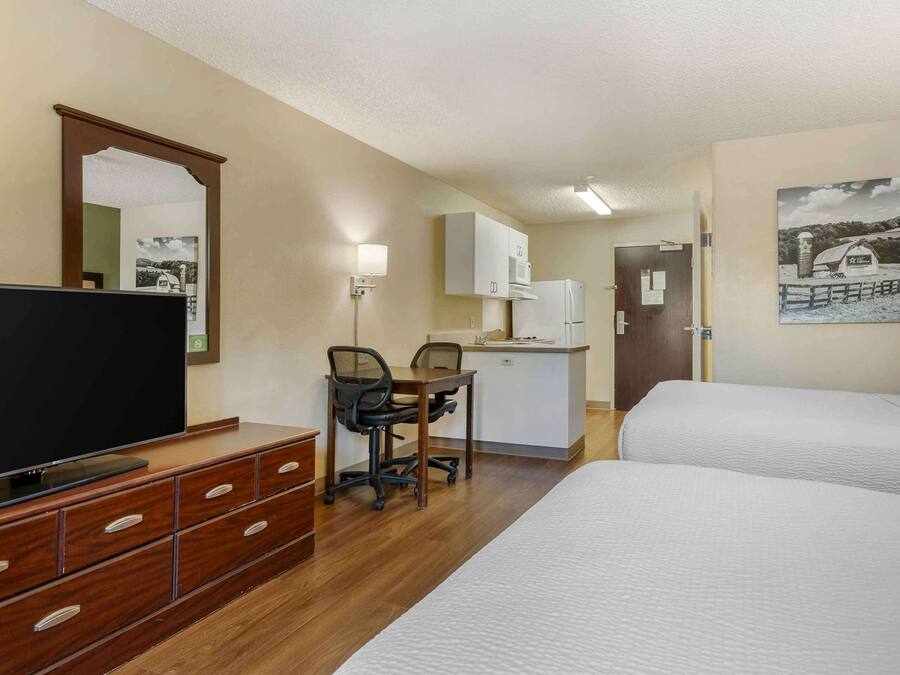A hotel room with a TV, wooden dresser, desk, two beds, kitchenette, and a painting on the wall.