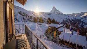 A snow-covered mountain with a sun shining behind it. There is a building with a balcony and outdoor seating.