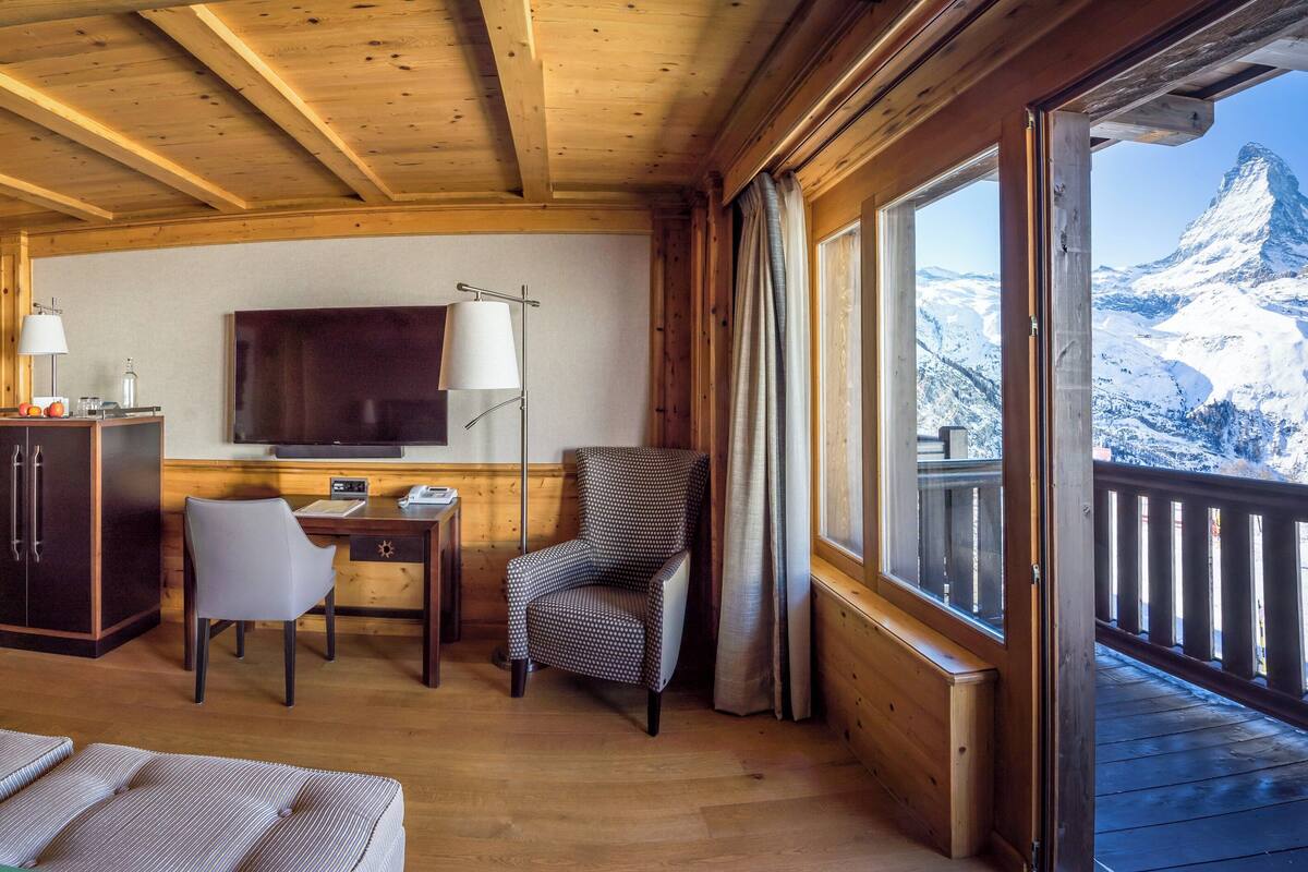 A cozy room with wooden walls and ceiling, a desk with a chair, a television, and a view of a snow-covered mountain.