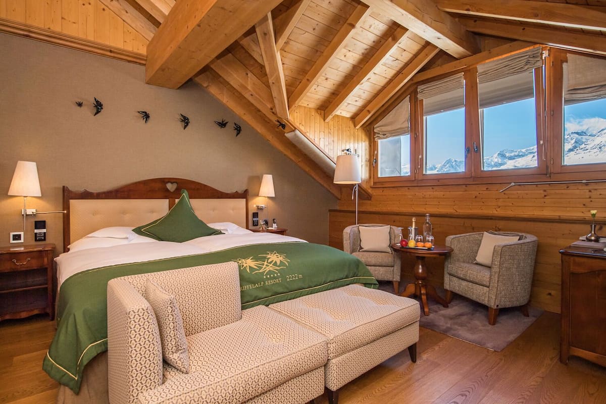 A cozy bedroom with a large bed, two bedside tables, a chair, a small table with drinks, and a window with a mountain view.